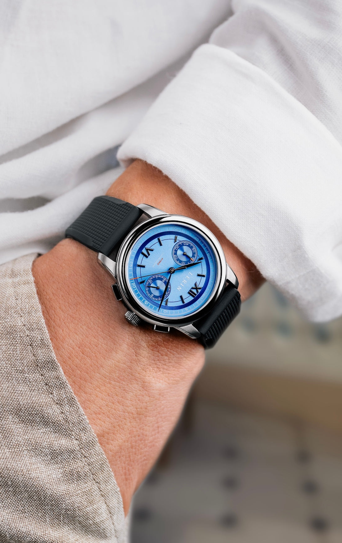 A close-up of a wrist wearing a silver-toned watch with a blue dial and black strap, shown on a person's wrist with a white shirt and beige trousers in the background.