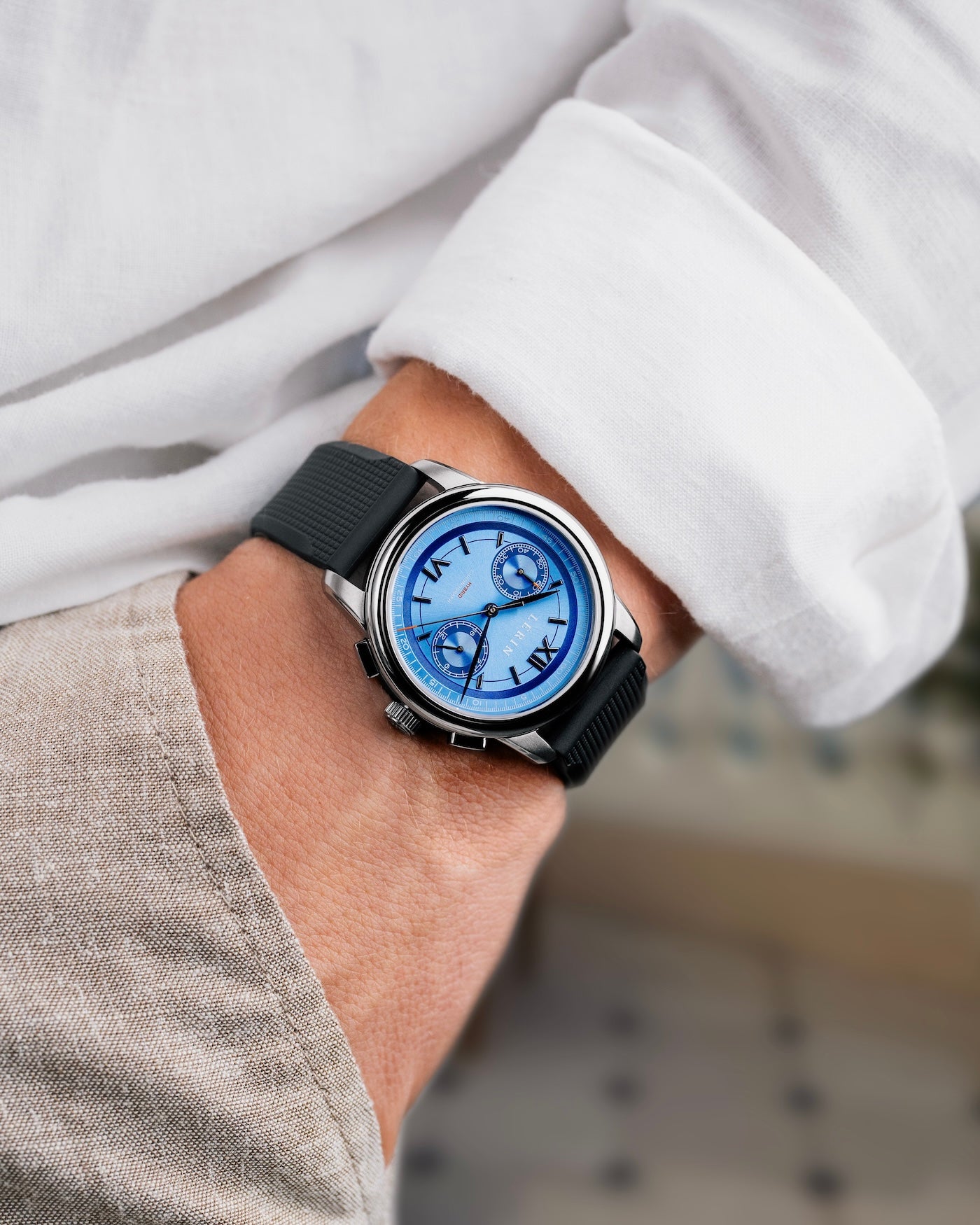 A close-up of a wrist wearing a silver-toned watch with a blue dial and black strap, shown on a person's wrist with a white shirt and beige trousers in the background.