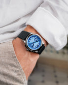 A close-up of a wrist wearing a silver-toned watch with a blue dial and black strap, shown on a person's wrist with a white shirt and beige trousers in the background.